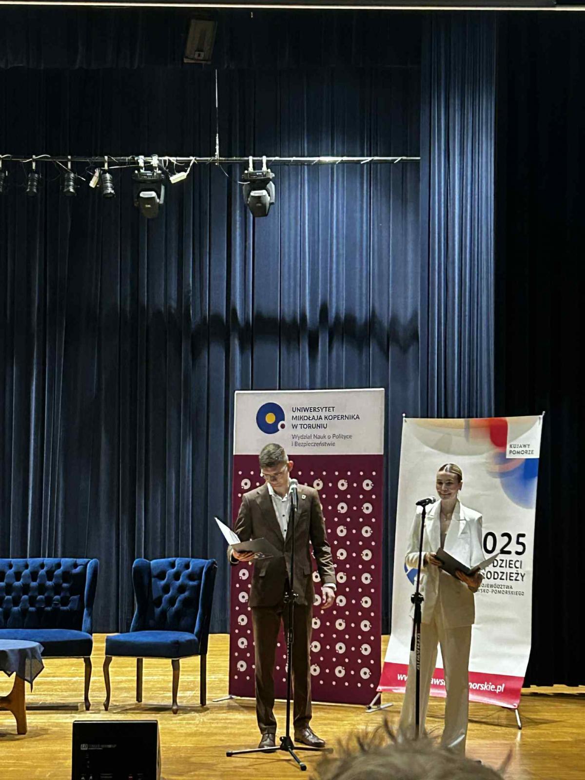 Dwie młode osoby, chłopak i dziewczyna, stoją na scenie przed mikrofonami