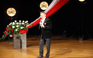 Jacek Wójcicki na scenie. W tle biało-czerwona flaga i bukiet róż.