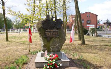Pomnik Niepodległości z wiązankami. Obok polskie flagi.