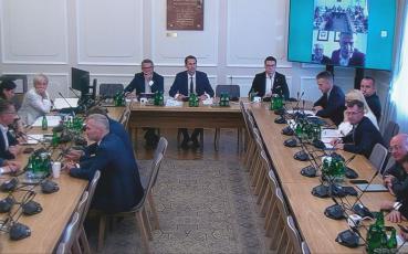 Zdjęcie sali posiedzeń. Za stołami siedzą członkowie komisji i zaproszeni goście. Na ekranie widać także osoby uczestniczące w posiedzeniu online.na 