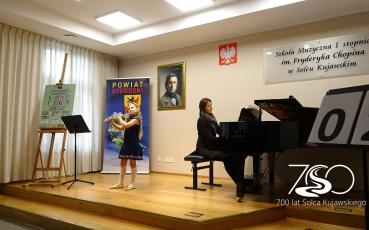 Występ młodej flecistki z akompaniatorem przy fortepianie. Na zdjęciu logo siedemsetlecia.
