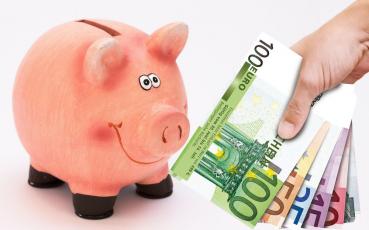 Różowa świnka - skarbonka patrzy uśmiechnięta na dłoń z banknotami euro.