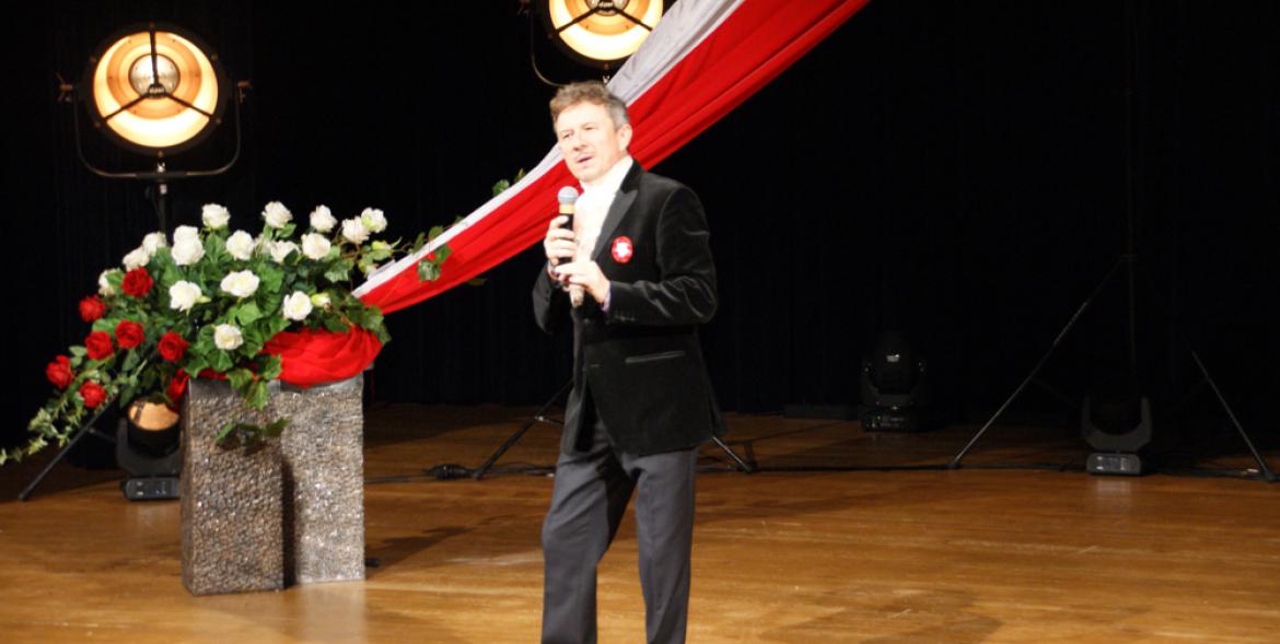 Jacek Wójcicki na scenie. W tle biało-czerwona flaga i bukiet róż.