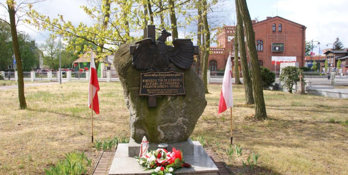 Pomnik Niepodległości, pod nim leżą wiązanki kwiatów, po jego obu stronach są biało-czerwone flagi