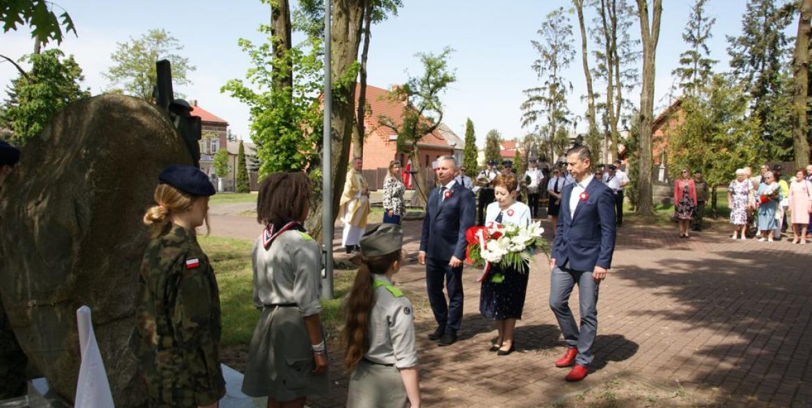 Delegacja z wiązanką kwiatów przed pomnikiem. Na pierwszym planie warta honorowa, w tle orkiestra OSP i pozostali uczestnicy uroczystości.