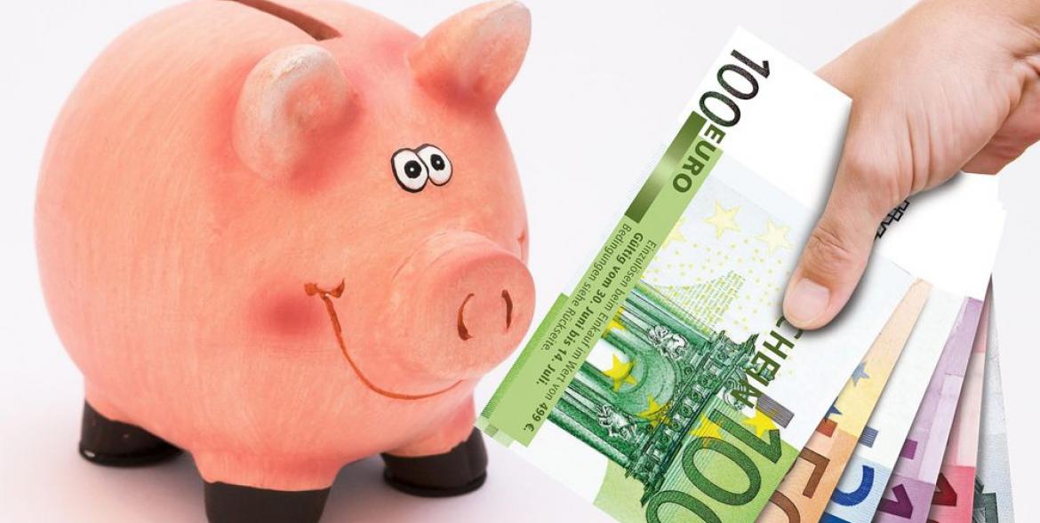 Różowa świnka - skarbonka patrzy uśmiechnięta na dłoń z banknotami euro.