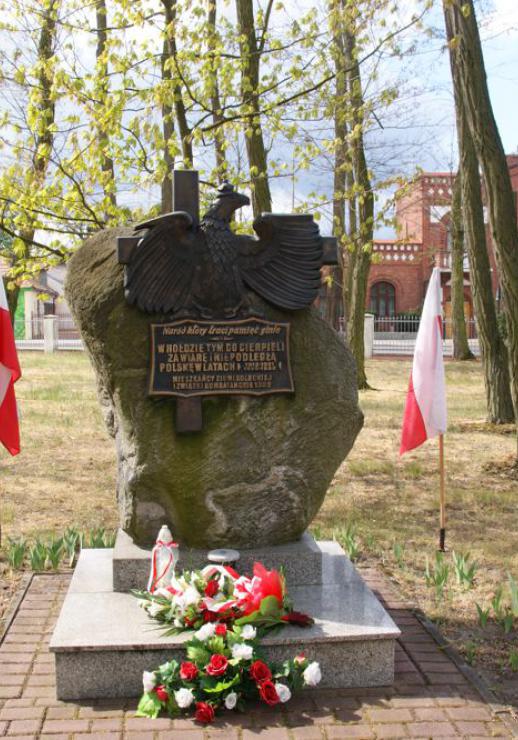 Pomnik Niepodległości z wiązankami. Obok polskie flagi.